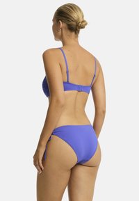 Eine Frau mit einem Dutt trägt einen lila Bikini, von hinten gesehen. Das Bikini-Oberteil hat dünne Träger und geraffte Details.