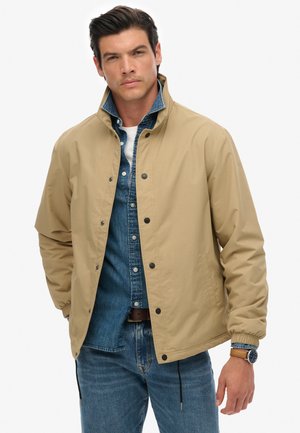 Lichte tan jas met hoge kraag, drukknoopsluiting, elastische manchetten en pockets, gelaagd over een spijkerhemd, gecombineerd met blauwe jeans.