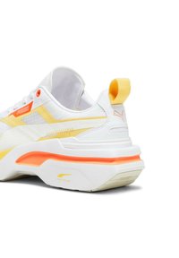 Baskets blanches avec des accents jaunes et oranges, tige en maille texturée, col rembourré et semelle en caoutchouc épaisse avec un design en couches.