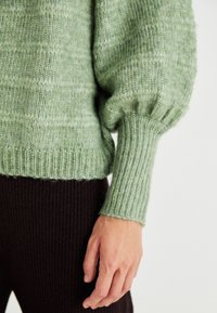 Knitted sweater in soft light green with textured pattern, wide sleeves, and ribbed cuffs. Paired with dark ribbed fabric.