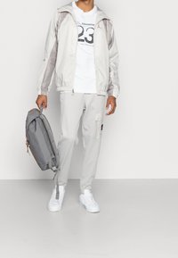 Ljust grå vindjacka med vita accenter, vit grafisk t-shirt, ljusgrå joggingbyxor och vita sneakers; grå ryggsäck med dragkedja.