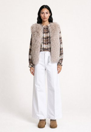 Woman standing wearing a beige fluffy vest, brown plaid shirt, wide white pants, and brown slip-on shoes against a plain light background.