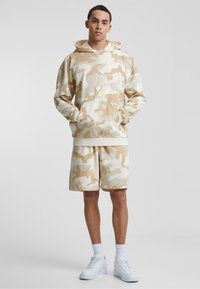 Beige camo hoodie with kangaroo pocket and matching shorts. Made of soft fabric; both pieces have a relaxed fit and subtle texture.
