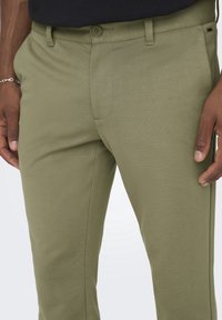 Pantalon vert en tissu texturé, avec fermeture à boutons et passants pour ceinture. Coupe slim, lignes épurées et détails de couture subtils.