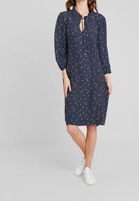 Tmavě modré šaty s volánkovým výstřihem, dlouhými rukávy a bílými puntíky. Má výřez ve tvaru klíče na přední straně a volný střih.