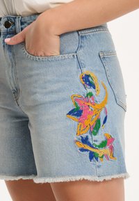 Gaiši zili džinsa šorti ar krāsainu izšūtu ziedu dizainu sānos, saplēstām malām un standarta kabatām.