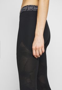 Czarne legginsy sportowe z teksturowanym siateczkowym panelem po boku, z elastycznym paskiem w wzór. Materiał wydaje się elastyczny i dopasowany.