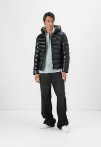 Schwarze Daunenjacke mit Kapuze, mit gestepptem Design und Reißverschluss. Kombiniert mit einem hellblauen Hemd und dunklen Jeans. Weißes Sneakers sichtbar.