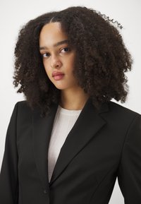 Black blazer with structured shoulders, tailored fit, and smooth texture. Worn over a light-colored ribbed top, showing a professional look.