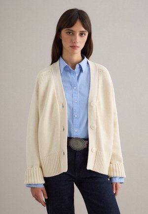 Giovane donna che indossa un cardigan lavorato a maglia color panna sopra una camicia a righe blu con pantaloni neri e fibbia decorativa, in piedi davanti a uno sfondo semplice.