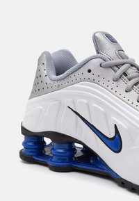 Nike Sportschuh mit einem Obermaterial in Weiß und Grau, silbernen Akzenten, blauen Dämpfungselementen und einem schwarzen Swoosh-Logo. Texturierter Bereich für die Schnürsenkel.