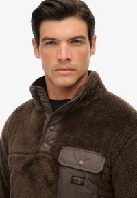 Brun fleece pullover med knapkrave, en tekstureret finish og en brystlomme med et mærke. Blød udseende og praktisk design.
