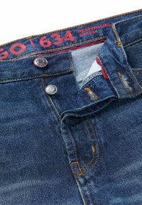 Jeans i denim i mörkblått med kopparstickning. Har två metallknappar i midjan och en röd etikett med vit text synlig inuti.