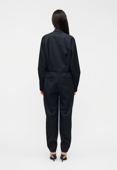 Σκούρο μπλε ολόσωμη φόρμα με κομψή σιλουέτα, μακριά μανίκια και εφαρμοστή μέση. Διαθέτει φερμουάρ και πλαϊνές τσέπες, κατασκευασμένη από υφή υφάσματος.