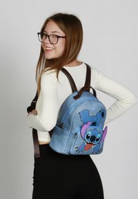 Mochila de color azul claro con diseño del personaje Stitch, correas marrones, bolsillos con cremallera y una textura suave.