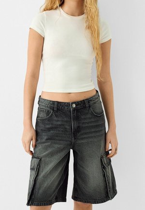 Person iført en tætsiddende hvid kortærmet crop top og løse, knælange sorte denim cargo shorts, stående foran en ensfarvet baggrund.