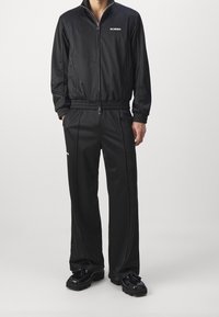 Black zip-up jacket and matching pants made from sleek, lightweight material. Features elastic cuffs and waistband, with subtle branding detail.