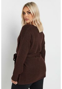 Brun stickad kofta med knyt i midjan, långa ärmar och avslappnad passform. Den texturerade tyget ger den ett mysigt utseende.