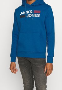 Modrá mikina s kapucí, s přední kapsou typu klokanka. Na přední straně je nápis "JACK & JONES" potištěný bílými a barevnými pruhy.