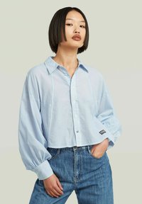 Camicia azzurra chiara, corta, con colletto classico, maniche a palloncino arricciate e un piccolo logo ricamato sull'orlo inferiore.
