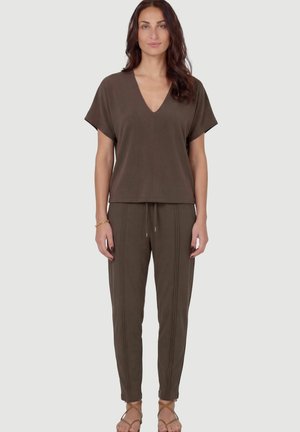Vrouw staand, draagt een bruin V-hals top met korte mouwen en bijpassende broek met trekkoord en verticale stiksels, bruine sandalen met bandjes en een gouden armband.