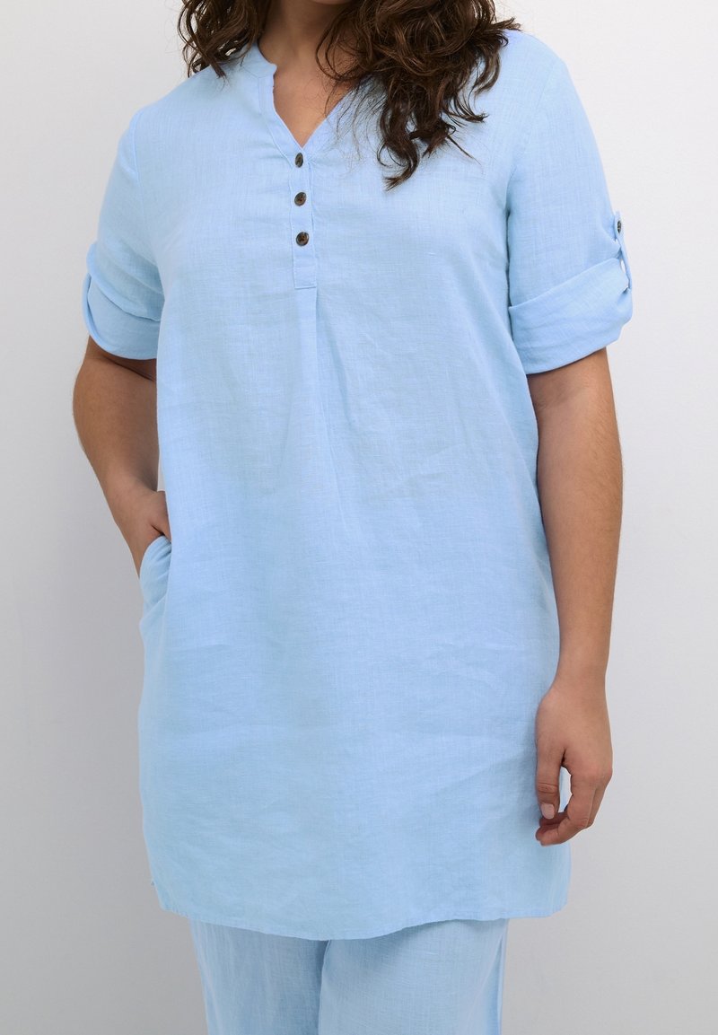 Tunique bleu clair avec des manches courtes roulées, patte de boutonnage et poches latérales. Fabriquée en tissu léger avec une texture lisse.