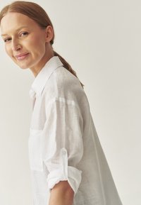 White linen shirt with a collar, rolled-up sleeves, and a chest pocket. Soft texture and lightweight appearance. Simple design with no patterns.