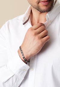 Närbild av en man i en vit skjorta som justerar sin krage, bärande ett silver- och svartmönstrat armband på sin vänstra handled.