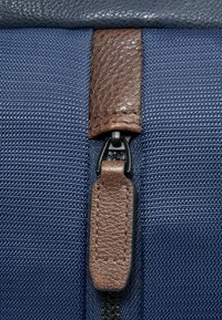 Marineblauwe, gestructureerde stof met een bruine leren accent en ritssluiting. Voorzien van een zwarte rits en nette stikdetails.