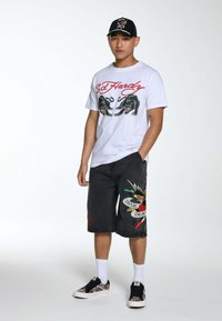 T-shirt bianco con grafica di pantera nera e testo rosso, pantaloni corti di denim neri con arte colorata in stile tatuaggio, scarpe da ginnastica nere con accenti a fantasia.
