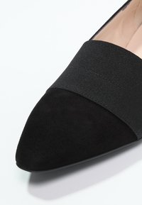 Chaussure plate en suède noir pointue avec large bride élastique sur le dessus et doublure intérieure beige clair visible.