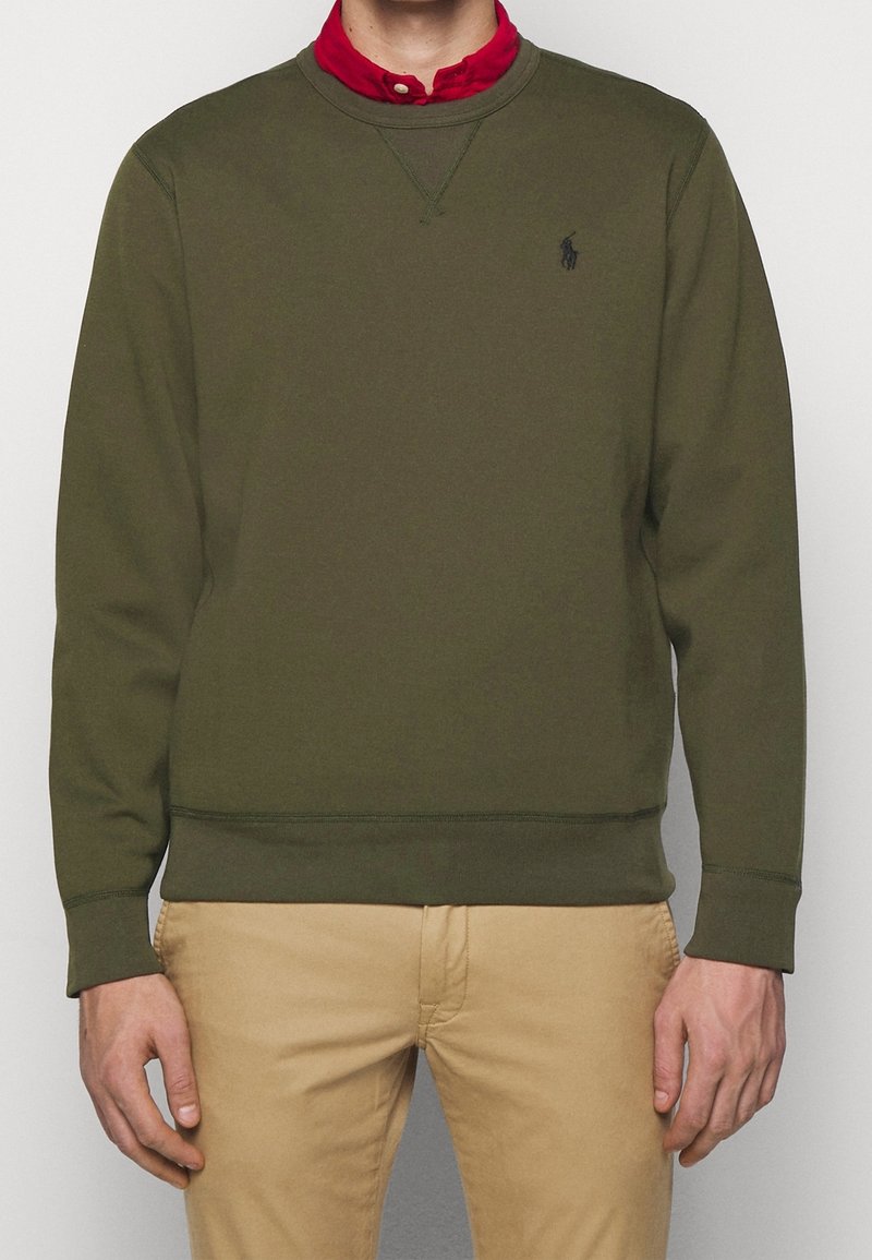 Olivgrön sweatshirt med rund halsringning, ribbade ärmslut och nederkant. Har en liten svart logotyp på bröstet. Lagerlagd över en röd skjorta med krage.