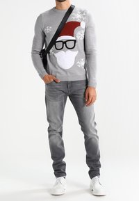 Mann in einem grauen Weihnachtspullover mit einem Santa-Gesicht, grauen Jeans, weißen Schuhen und einer schwarzen Umhängetasche, steht mit einer Hand in der Tasche.