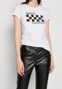 Bílý bavlněný tričko s černým kostkovaným vzorem a textem "ARMANI EXCHANGE". Krátké rukávy a klasický design s kulatým výstřihem.