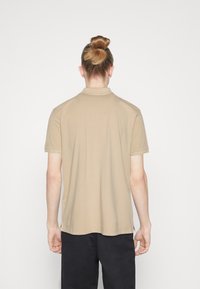 Polo beige à manches courtes avec une coupe droite, une texture douce et des lignes épurées ; il présente un col classique et de discrètes fentes latérales.