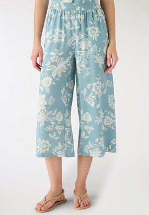 Pantaloni larghi color azzurro chiaro a gamba larga e taglio corto con stampa floreale bianca, indossati con sandali infradito beige con borchie, mostrati dalla vita ai piedi.