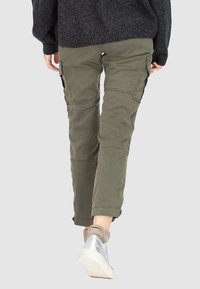 Olivově zelené cargo kalhoty s zužujícím se střihem, bočními kapsami a odolnou tkaninou. Nošeny se šedou obuví a černým pleteným svetrem.