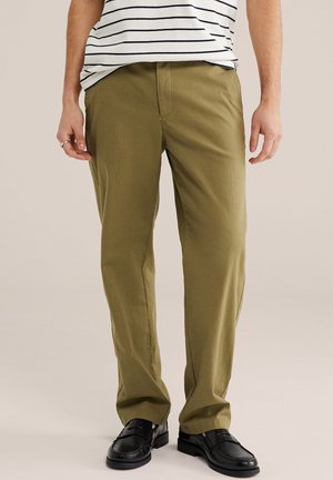 Homme portant un pantalon vert olive, des mocassins en cuir noir et une chemise blanche à rayures horizontales noires, debout devant un fond neutre.