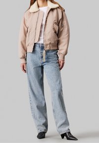 Jasno beżowa kurtka bomberka z kołnierzem z owczej skóry, zapinana na zamek, z bocznymi kieszeniami, w połączeniu z jasnoniebieskimi, leżącymi w talii dżinsami oraz czarnymi botkami na obcasie.