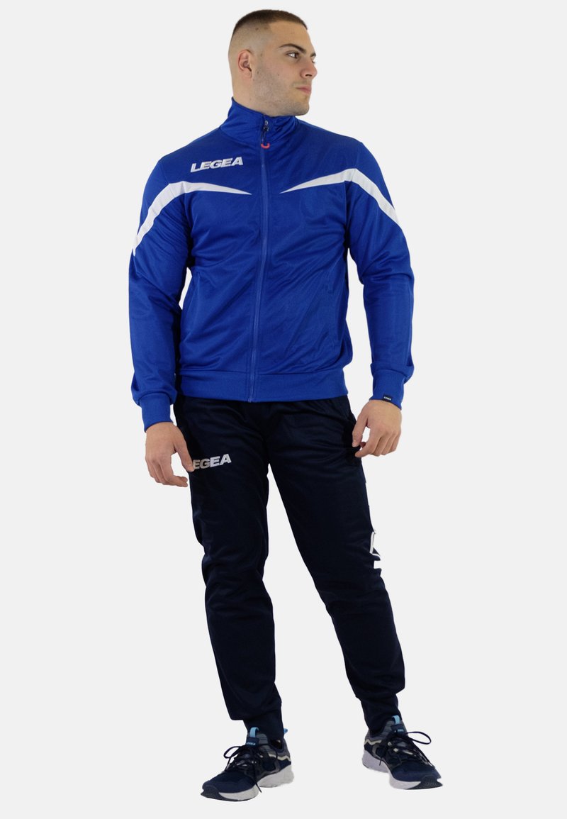 Blå atletisk jakke med hvide accenter, lynlåsefront, parret med sorte bukser. Har branding på jakken og bukserne, glat tekstur.