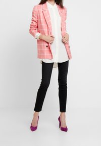 Blazer a cuadros rosa con botones negros, camisa blanca debajo, jeans negros ajustados y tacones altos morados. Textura suave y corte a medida.