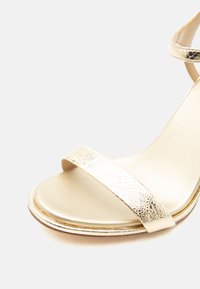 Κοντινή λήψη χρυσού μεταλλικού πέδιλου με ανοιχτά δάχτυλα, με έναν μόνο μπροστινό λουράκι και λουράκι στο αστράγαλο, σε απλό ανοιχτό φόντο.