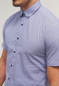 Košile s krátkým rukávem v modro-bílém kostkovaném vzoru. Má límec na knoflíky a tmavé knoflíky, je vyrobená z lehké látky.