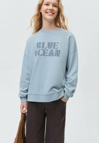 Svetlomodrý sveter s grafikou "BLUE OCEAN" v tmavosivej farbe. Obsahuje voľný strih, okrúhly výstrih a rebrované manžety. Nosí sa s tmavými nohavicami.