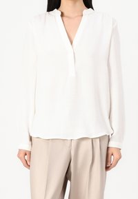 Bílá blůza s dlouhými rukávy, véčkovým výstřihem a vlnitým límečkem. Měkká, lehká látka s volným střihem, spárovaná s béžovými kalhotami.
