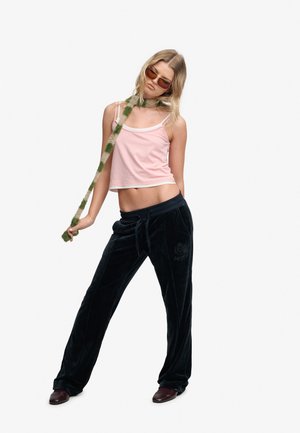 Ung kvinde iført en lyserød cropped tanktop, sorte fløjlsbukser, solbriller og et dunet grønt og beige tørklæde med hænderne i lommerne.