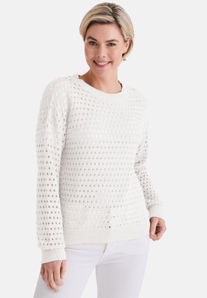 Lächelnde Frau mit kurzen blonden Haaren, die einen weißen, langärmeligen, strukturierten Strickpullover und weiße Hosen vor einem schlichten Hintergrund trägt.