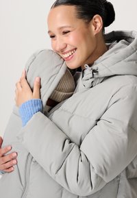 Een vrouw die warm glimlacht terwijl ze een baby in een muts omarmt, beiden in lichtgrijze gewatteerde jassen, tegen een neutrale achtergrond.