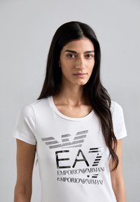 T-shirt bianca in cotone con scollatura tonda, caratterizzata da una stampa in rilievo nera del logo "EA7 Emporio Armani" sulla parte anteriore.