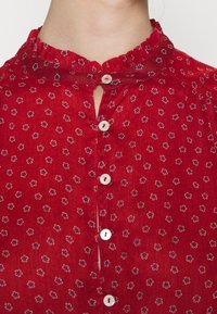 Punainen pusero, jossa on röyhelökaulus, avainaukko-aukko ja valkoiset nappisulkimet. Koristeena mustavalkoinen kukkakuosi. Pehmeä tekstuuri.